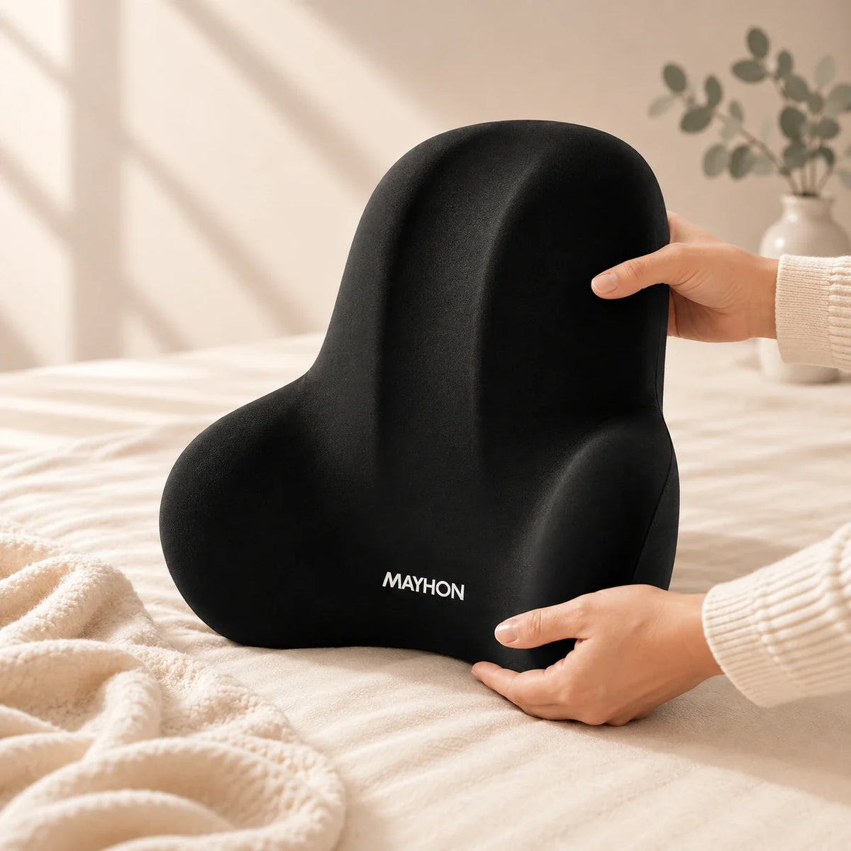 Coussin lombaire noir à contours ergonomiques, tenu à deux mains sur un lit. Image